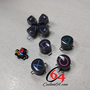 May include: A set of black gaming controller replacement parts including a directional pad, four round action buttons with blue X, pink O, green triangle, and purple square symbols, and a small black and multicoloured logo emblem with Custom64.com text.