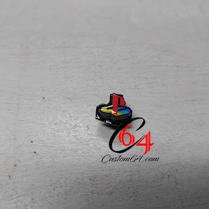 Puede incluir: Un pequeño pin de esmalte metálico con el icónico logotipo de PlayStation en colores rojo, amarillo, verde y azul sobre un fondo negro. El pin está colocado sobre un gráfico de logotipo negro y rojo que incluye el texto Custom64.com.