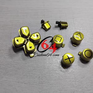 May include: A collection of gold-colored controller buttons and directional pad. The buttons feature symbols like a square, triangle, circle, and cross. The directional pad has an arrow design. The text "Custom64.com" is also visible.