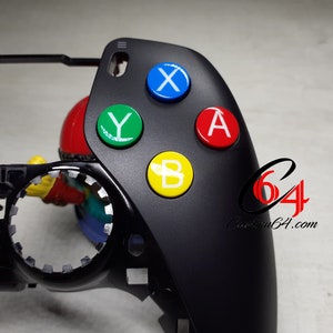 Può includere: Controller per videogiochi nero con quattro pulsanti colorati: blu, verde, giallo e rosso. I pulsanti sono etichettati con X, Y, B e A. Al controller manca la placca frontale.