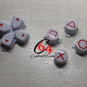May include: Set of six white plastic video game controller buttons with red symbols. The buttons feature the symbols: triangle, circle, X, square, and two arrows.