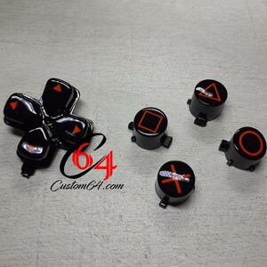 May include: Black and orange PlayStation controller buttons. The buttons are shaped like a cross, a circle, a square, a triangle, and an X. The buttons are on a gray surface.