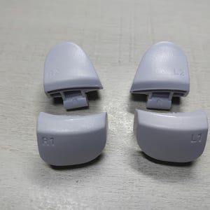 Peut inclure: Quatre boutons de contrôleur gris clair, dont deux gâchettes marquées "R2" et "L2", et deux boutons d'épaule marqués "R1" et "L1". Les boutons sont en plastique et sont destinés à une manette de jeu.