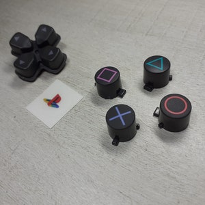 May include: A set of six black replacement buttons for a PlayStation controller. The buttons feature different coloured symbols: a pink square, a blue triangle, a red circle, a blue X, and two grey arrows.