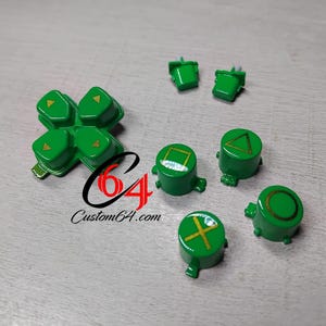 Puede incluir: Botones de repuesto verdes para un mando de juegos, incluyendo una cruceta y botones individuales con símbolos dorados. Los botones están dispuestos sobre una superficie gris claro. El sitio web Custom64.com es visible.