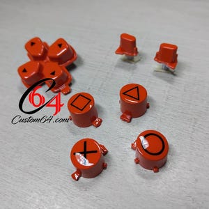 May include: Orange replacement gaming console buttons, including a directional pad, triangle, square, circle, and cross symbols. The buttons are displayed on a light grey surface. The Custom64.com logo is also visible.