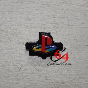 Puede incluir: Botón negro de PlayStation con el icónico logotipo rojo, amarillo, azul y verde. El botón tiene un diseño en forma de cruz y el texto "Custom64.com" es visible.