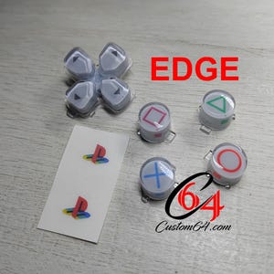 Peut inclure: Un ensemble de boutons de manette PlayStation blancs avec différents symboles colorés, dont un carré rouge, un triangle vert, un cercle bleu et un X bleu. Les boutons sont étiquetés "EDGE". Il y a aussi deux autocollants avec le logo PlayStation.
