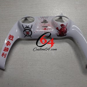 Może przedstawiać: Biała plastikowa płytka czołowa do kontrolera gier wideo z czerwonymi i czarnymi japońskimi znakami i wzorami. Płytka czołowa ma dwa otwory na przyciski kontrolera i efekt rozpryskanej farby.