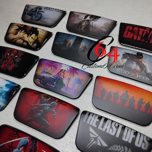 Puede incluir: Una colección de accesorios rectangulares con varios gráficos de la cultura pop. Los diseños incluyen Batman, The Last of Us y otros personajes de videojuegos y cómics. Los accesorios tienen un acabado brillante y están dispuestos sobre una superficie clara.