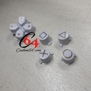 May include: Set of six white plastic buttons with gold symbols. The symbols are a circle, a cross, a square, a triangle, and two arrows.