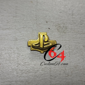 Può includere: Uno spillo con il logo PlayStation dorato. Il logo è in rilievo e ha un effetto 3D. Lo spillo è su uno sfondo bianco.
