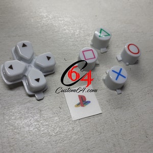 May include: White replacement buttons for a PlayStation controller. The buttons feature the standard PlayStation symbols: circle, cross, triangle, and square. The buttons are arranged in a row, with the PlayStation logo in the center.