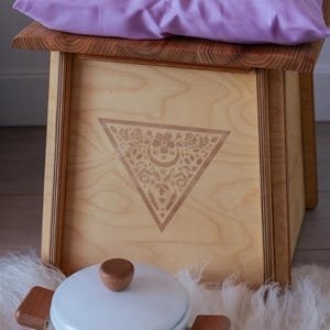 a wooden stool with a purple pillow on top, placed on a white furry surface. Next to the stool is a white ceramic pot with a wooden handle.