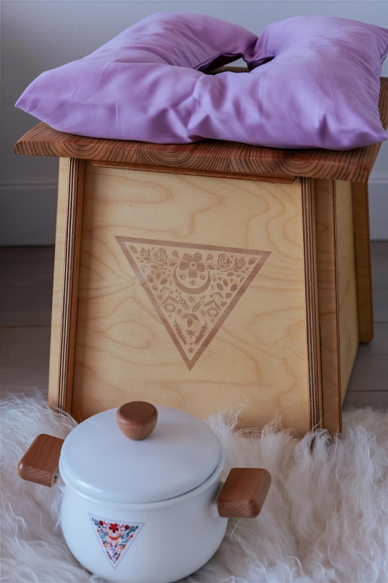 a wooden stool with a purple pillow on top, placed on a white furry surface. Next to the stool is a white ceramic pot with a wooden handle.