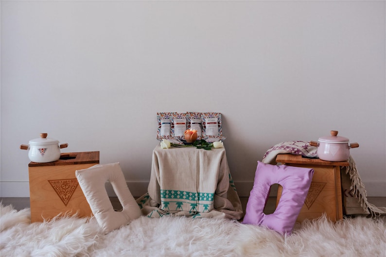 a cozy and inviting scene with a white rug, wooden crates, and various decorative items. There are two white pots, one on each side of the image, and a wooden crate with a pink pot on top. A wooden table is also present, with a vase and a candle placed on it. The arrangement of the objects creates a warm and welcoming atmosphere.