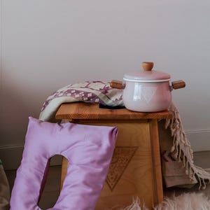 a wooden stool with a pink pot on top, placed on a fluffy white rug. There is also a purple pillow and a blanket on the stool.