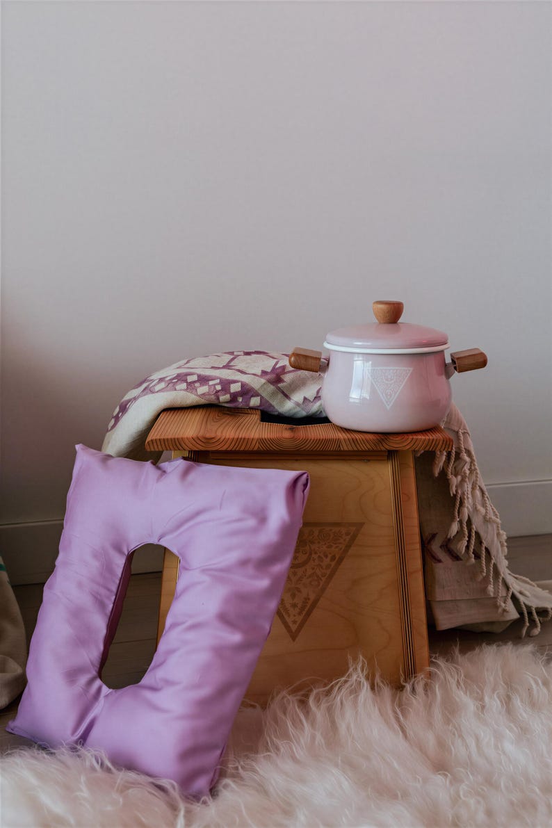 a wooden stool with a pink pot on top, placed on a fluffy white rug. There is also a purple pillow and a blanket on the stool.