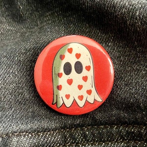 May include: A round, red button featuring a cartoon ghost with heart-shaped eyes and accents. The ghost is white with a green outline and red hearts. The button is set against a dark denim background.