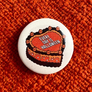 May include: A white button with a brown and orange heart-shaped cake with the text "Every Day is Halloween" on it.