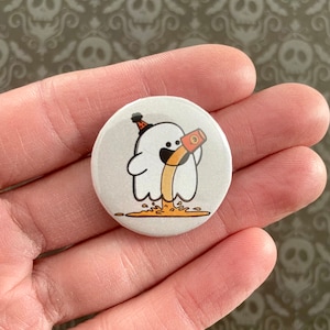 May include: A round, white button featuring a cartoon ghost pouring a beverage from a brown cup. The ghost wears a party hat. The beverage is orange, and the button has a glossy finish.