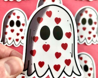 May include: White ghost sticker with red heart accents. The ghost has large black eyes and a scalloped bottom edge. Several similar stickers are visible against a pink background. The sticker is held by a hand.
