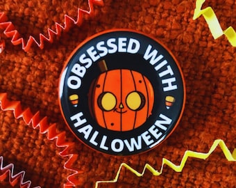May include: A round, black button with the words "OBSESSED WITH HALLOWEEN" in white. The button features a cartoon jack-o'-lantern with a smiling face. Orange and yellow decorative paper is scattered around the button on an orange textured surface.