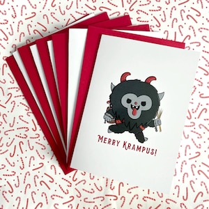 May include: A stack of greeting cards with red and white accents. The front card features a cartoon Krampus character with the text "MERRY KRAMPUS!". The background is a pattern of red and white candy canes.