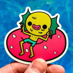 May include: A yellow cartoon character sticker with a green fin-like head, wearing teal shorts, and holding a drink. The character is on a pink floatie with white ghost-like shapes. The sticker has a white border.