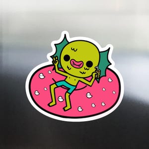 May include: A sticker featuring a cartoon creature with a green head, blue shorts, and a pink inner tube. The creature is holding a drink and giving a peace sign. White ghost illustrations are scattered on the pink tube.