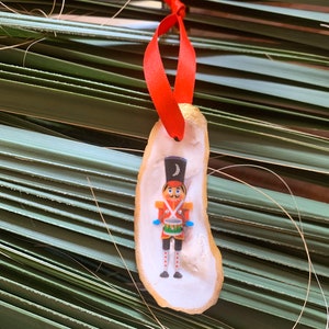 May include: A red and gold Christmas ornament featuring a nutcracker soldier holding a drum. The ornament is made from a natural oyster shell.