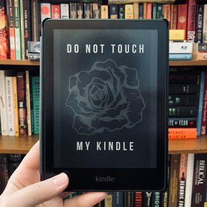 May include: Black Kindle e-reader with the text "DO NOT TOUCH MY KINDLE" and a rose design on the screen. The e-reader is held in front of a bookshelf filled with books.