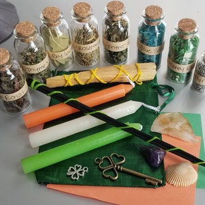 May include: A green pouch with a variety of herbs, crystals, and candles. The herbs are in small glass bottles with cork tops and labels that read "Basil", "Thyme", "Bay Leaf", "Rosemary", "Mint", "Blue Salt", "Money Bag", and "Pyrite". The pouch also contains a white candle, a green candle, an orange candle, a black stick, a small silver key with a clover charm, a small amethyst crystal, a small clear quartz crystal, and a seashell.