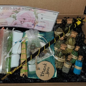 May include: A black box filled with black shredded paper and various items, including a small wooden stick, a white cloth bag, a small white ceramic ring, a small glass bottle with a cork stopper, and a small piece of paper with the words "thank you" written on it. The box also contains a check with the words "The Universal Bank of Abundance" printed on it.