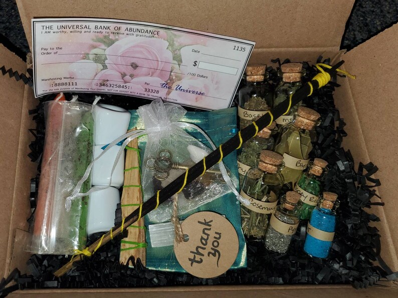 May include: A black box filled with black shredded paper and various items, including a small wooden stick, a white cloth bag, a small white ceramic ring, a small glass bottle with a cork stopper, and a small piece of paper with the words "thank you" written on it. The box also contains a check with the words "The Universal Bank of Abundance" printed on it.