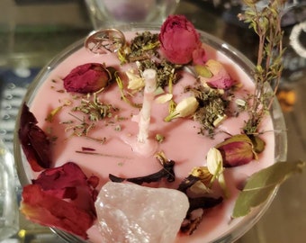 Pode incluir: Vela rosa com flores secas, ervas e um cristal de quartzo transparente. A vela está em um frasco de vidro com um amuleto prateado na borda. O texto no frasco diz "Aumente sua capacidade psíquica".