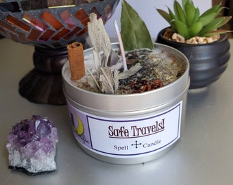 May include: A silver candle tin with a white label that reads "Safe Travels! Spell + Candle". The candle is filled with herbs, dried flowers, and a cinnamon stick.