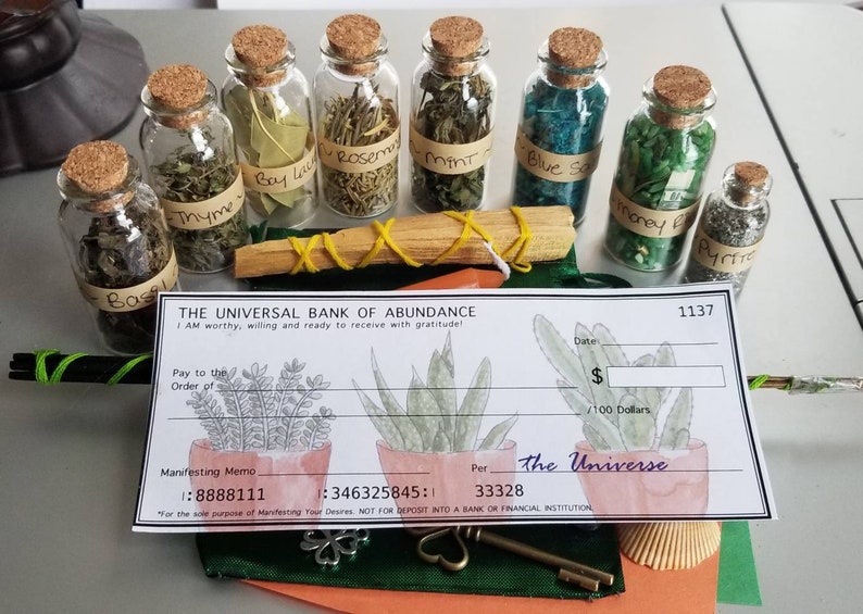 May include: A set of small glass bottles with cork tops filled with dried herbs and crystals. The bottles are labeled with the names of the herbs and crystals, including Basil, Bay Leaf, Rosemary, Thyme, Mint, Blue Salt, Money Bag, and Pyrite. The bottles are arranged in a row on a table, along with a wooden stick and a check printed with "The Universal Bank of Abundance".