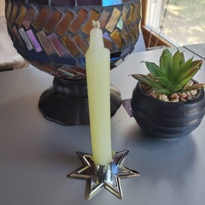 May include: A yellow candle sits in a silver star-shaped candle holder. The candle is tall and thin, and the holder has a simple design.