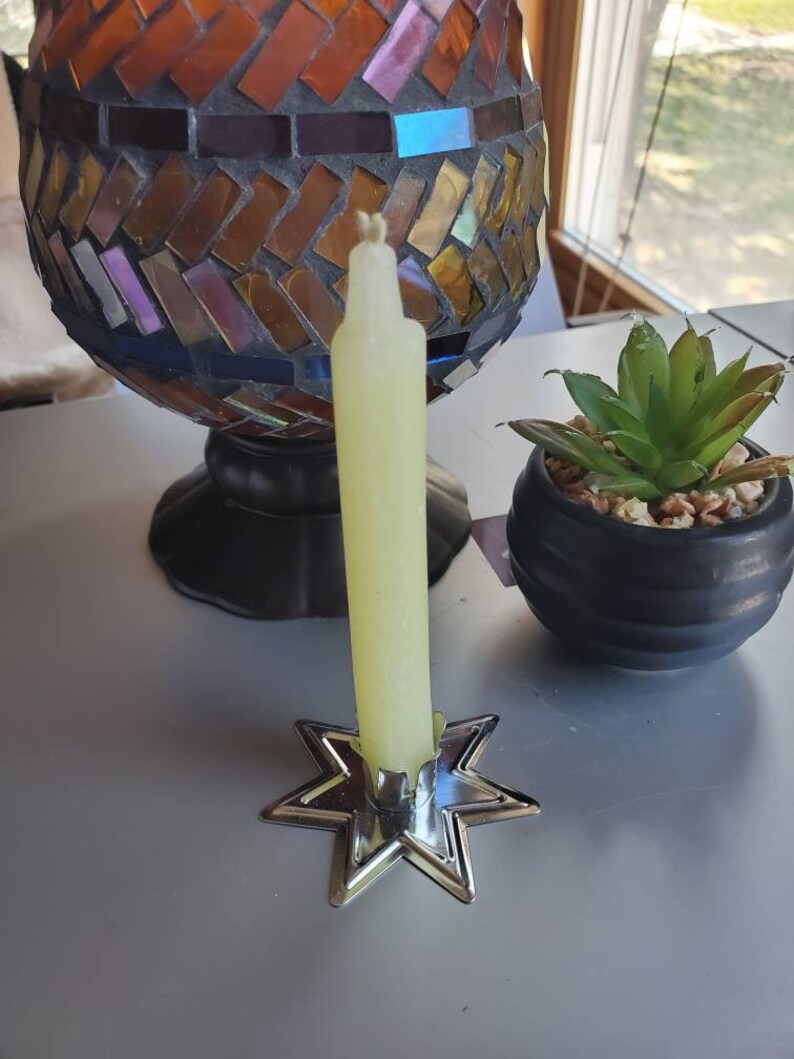May include: A yellow candle sits in a silver star-shaped candle holder. The candle is tall and thin, and the holder has a simple design.
