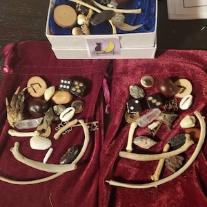May include: Two red velvet bags filled with various objects, including bones, crystals, shells, dice, and runes. The bags are sitting on a white surface next to a white box with a blue velvet interior. The box contains similar objects and a logo that says "Spellcraft".