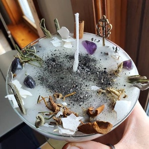 May include: A white candle with a central wick, adorned with amethyst crystals, dried herbs, eggshell fragments, and a decorative key charm. The candle has a glass base and is sprinkled with black powder.
