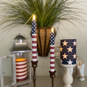 May include: Two lit, patriotic-themed candles with a blue star-studded top and red and white stripes. The candles are in bronze candle holders. A remote control sits in front of the candles. A matching star-patterned lamp and a lantern are also visible.
