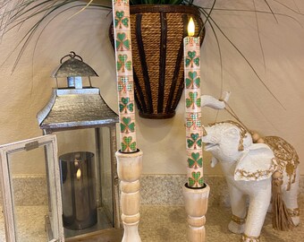 May include: Two battery-operated candles with shamrock patterns are in light-coloured wooden candle holders. A remote control is in front of the candles. A decorative lantern and a white elephant statue are also in the image.