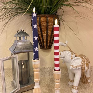 May include: Two decorative candles with patriotic designs, one with a blue background and white stars, the other with red and white stripes. They are placed on wooden candle holders. A white elephant statue and a lantern are also in the image.
