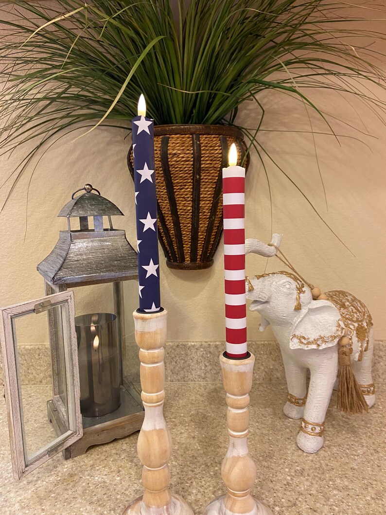 May include: Two decorative candles with patriotic designs, one with a blue background and white stars, the other with red and white stripes. They are placed on wooden candle holders. A white elephant statue and a lantern are also in the image.