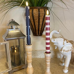 May include: Two decorative candles in wooden holders. One is navy blue with white stars, the other red and white striped. A lantern and elephant figurine are also visible. The candles appear to be battery operated.