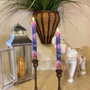 May include: Two purple Halloween candles with pumpkin and bat designs, set in bronze candle holders. A decorative lantern and a white elephant statue are in the background, along with a hanging basket of greenery.