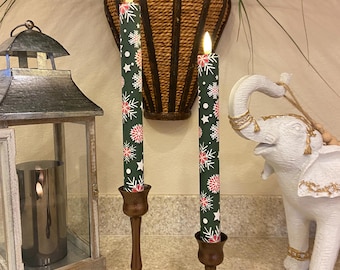 May include: Two battery-operated Christmas candles with a green background and red and white snowflake design. The candles are in bronze-colored candle holders. A remote control is in front of the candles. A decorative elephant statue is to the right.