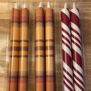 May include: Candles in clear plastic packaging. Two sets of candles are visible. One set has an orange plaid pattern, and the other has red and white stripes. The text "Example of semi-gloss finish" is above the candles.